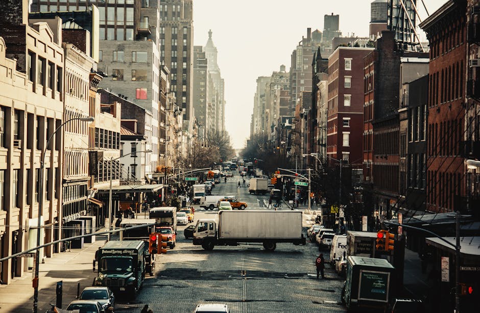 A city street scene of a busy urban area with tall buildings on both sides, featuring a range of vehicles including trucks, vans, and cars moving along the road. The street is lined with commercial and residential structures, with some storefronts visible at street level. Several delivery and moving trucks are stationary or moving through the traffic, and a few pedestrians can be seen crossing or walking along the pavements. The lighting suggests late afternoon with soft shadows cast by the buildings, and the overall scene captures typical city traffic and activity, relevant to house removals and furniture transport services like Chelsea Movers conducting packing, loading, and home relocation processes in the area near Kings Road flats, Chelsea.