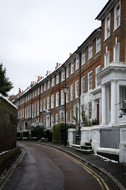 A row of red-brick terraced townhouses with white window frames and decorative columns at the entrance, situated along a narrow, slightly curved street in SW3. The pavement is bordered by a low brick wall and greenery, with some leafless shrubs and trees. Outside one of the homes, there are cardboard boxes, wrapped furniture, and plastic-wrapped household items placed on the front steps and along the sidewalk, indicating a house move in progress. A moving trolley and packing materials are also visible near the entrance, suggesting the ongoing packing and loading process. The scene is captured in natural daylight with overcast skies, providing a clear view of the exterior environment typical of Chelsea residential areas. Chelsea Movers are likely involved in the home relocation and furniture transport services for the property, supporting the detailed logistics associated with removals for SW3 townhouses.