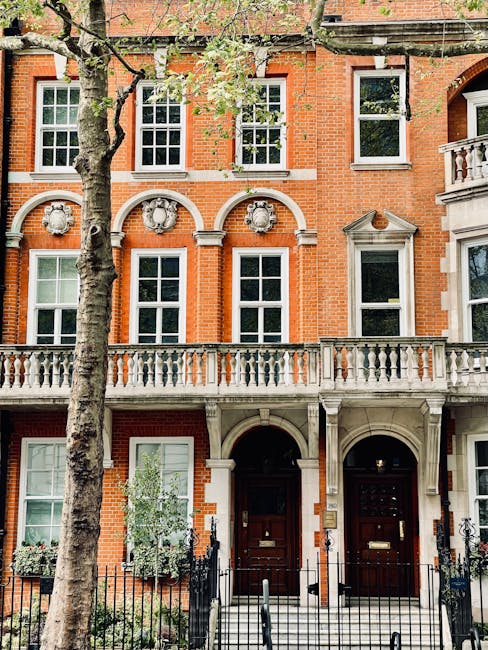 An exterior view of a Georgian-style red brick townhouse with three large white-framed sash windows on the upper floors, and two black wooden doors on the ground level, each with a brass letterbox and door knocker. The building features decorative stone arches above the front doors and a small balcony with a balustrade on the right side. In front of the property, a paved area is enclosed by a low black wrought-iron fence. A leafless tree partially obscures the facade, and green potted plants are positioned on window sills and near the entrances. This scene depicts a residential property in an urban area, suitable for house removals or home relocation services, with natural daylight highlighting the architectural details. Chelsea Movers, specializing in removals, might be involved in a furniture transport or packing and moving process associated with this type of townhouse.