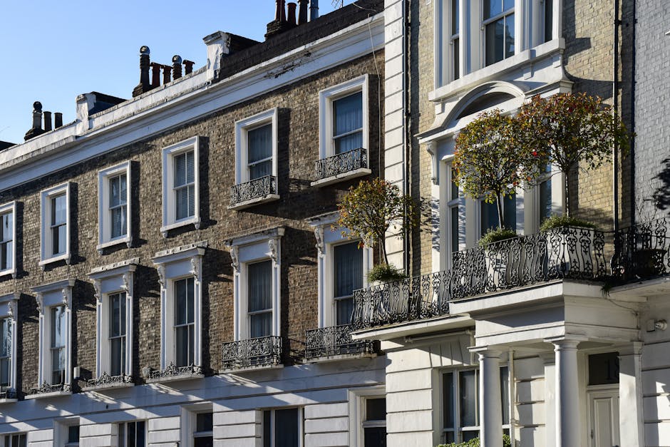 A row of red-brick terraced townhouses with white window frames and decorative columns at the entrance, situated along a narrow, slightly curved street in SW3. The pavement is bordered by a low brick wall and greenery, with some leafless shrubs and trees. Outside one of the homes, there are cardboard boxes, wrapped furniture, and plastic-wrapped household items placed on the front steps and along the sidewalk, indicating a house move in progress. A moving trolley and packing materials are also visible near the entrance, suggesting the ongoing packing and loading process. The scene is captured in natural daylight with overcast skies, providing a clear view of the exterior environment typical of Chelsea residential areas. Chelsea Movers are likely involved in the home relocation and furniture transport services for the property, supporting the detailed logistics associated with removals for SW3 townhouses.