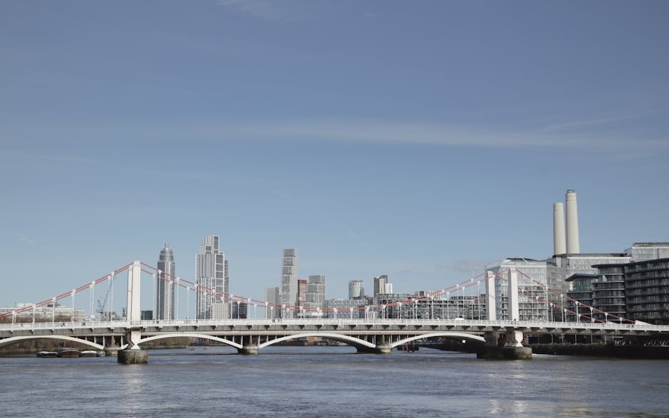 A panoramic view of a city skyline with several high-rise buildings, including one with a sleek modern glass facade, and two large industrial chimney stacks on the right. In the foreground, a river re