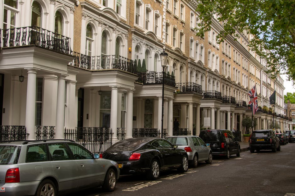 The image shows a row of elegant, Victorian-style terraced houses in Sloane Square, Chelsea, with white facades, decorative columns, and black wrought iron balconies. Several cars, including a silver estate, a black sports car, and dark-colored sedans, are parked along the street on the pavement adjacent to the properties. A black lamppost stands among the parked vehicles, and flags are visible hanging from some of the houses. Green trees partially shade the scene, indicating a bright day. The overall setting is indicative of a residential area where home relocation or furniture transport may take place, with the street prepared for moving logistics coordinated by Chelsea Movers, a professional removals company specializing in house moves in the Chelsea area, as suggested by the webpage context.