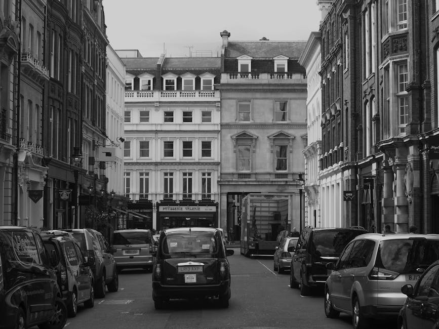 A narrow street scene in Chelsea, London, featuring multiple parked cars including small hatchbacks and vans along both sides of the road. Historic multi-storey buildings with ornate facades, large windows, and decorative architectural details line the street. In the background, a delivery van or moving truck is parked close to a building entrance, indicating a home relocation or furniture transport process. The pavement is visible, and the street appears to be in an urban area with limited parking space, relevant for house removals and parking tips for movers. The overall scene is captured in black and white, emphasizing the detail of the vehicles and surrounding architecture, which are common in urban moving contexts, with Chelsea Movers potentially assisting with such relocations.