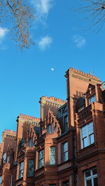 The image shows a row of elegant, Victorian-style terraced houses in Sloane Square, Chelsea, with white facades, decorative columns, and black wrought iron balconies. Several cars, including a silver estate, a black sports car, and dark-colored sedans, are parked along the street on the pavement adjacent to the properties. A black lamppost stands among the parked vehicles, and flags are visible hanging from some of the houses. Green trees partially shade the scene, indicating a bright day. The overall setting is indicative of a residential area where home relocation or furniture transport may take place, with the street prepared for moving logistics coordinated by Chelsea Movers, a professional removals company specializing in house moves in the Chelsea area, as suggested by the webpage context.